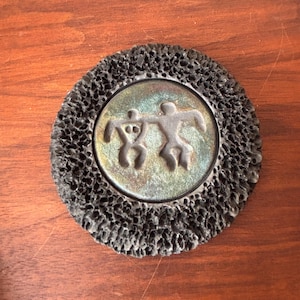 May include: A round, decorative object with a textured, dark gray outer ring. The center features a metallic, iridescent circle with two stylized human figures. The figures are in a dancing or celebratory pose.