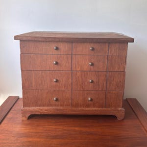 May include: A small wooden chest of drawers with eight drawers, each with a small, round knob. The chest is made of reddish-brown wood and has a simple, rectangular design. The chest sits on a dark wooden surface.