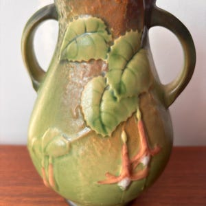 May include: Green ceramic vase with two handles, featuring embossed leaves and flowers in shades of green, orange, and white. The vase has a textured surface and a brown rim.