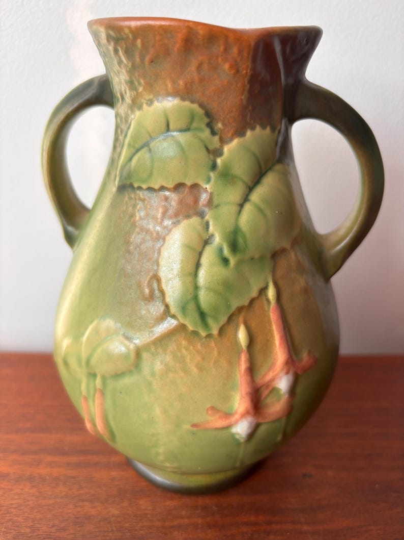 May include: Green ceramic vase with two handles, featuring embossed leaves and flowers in shades of green, orange, and white. The vase has a textured surface and a brown rim.
