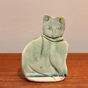 Könnte beinhalten: Eine Keramik-Katzenfigur in sitzender Position, glasiert in Grün- und Blautönen. Die Katze hat spitze Ohren, detaillierte Augen und einen Kragen. Der Sockel ist weiß. Die Figur steht auf einer Holzoberfläche.