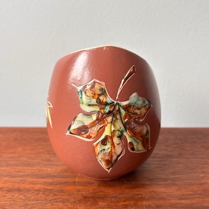 Puede incluir: Un pequeño jarrón de cerámica de color terracota con un diseño de hoja. La hoja está pintada con tonos de marrón, amarillo y verde. El jarrón tiene una forma redondeada y una superficie lisa. El jarrón está sobre una superficie de madera.