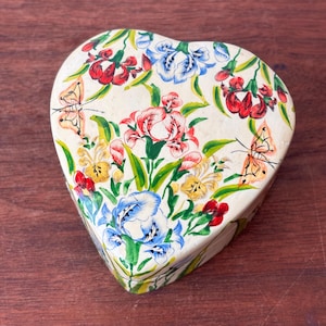 May include: Heart-shaped decorative box with a cream background. The box is decorated with a floral pattern of red, pink, blue, and yellow flowers, green leaves, and orange butterflies. It rests on a wooden surface.