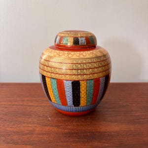 Może przedstawiać: Ozdobny słoik z pokrywką o żywym, kolorowym wzorze. Słoik ma czerwoną podstawę i pokrywkę, ze złotymi akcentami i kwiatowymi wzorami. Korpus słoika zdobią pionowe paski w różnych kolorach, w tym niebieskim, żółtym i czarnym.