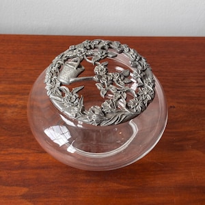 Pode incluir: Uma tigela de vidro transparente com uma tampa decorativa prateada. A tampa apresenta um design floral com um regador e flores. A tigela está sobre uma superfície de madeira. A tigela é redonda e tem uma abertura larga.