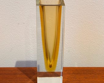 May include: A rectangular glass vase with a clear exterior and a golden-yellow interior. The vase has a square opening and a tapered design, creating a striking visual contrast. The vase is displayed on a wooden surface.