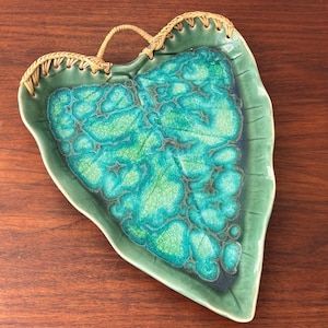 May include: Green leaf-shaped ceramic serving dish with a turquoise and green crackle glaze. The rim is stitched with a woven tan material, and it has a woven handle. The dish is on a wooden surface.