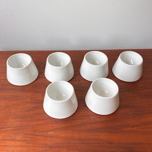 May include: Six white ceramic egg cups arranged on a wooden surface. Each cup has a rounded, open top and a gently sloping, conical shape. The cups are evenly spaced, showcasing their minimalist design.