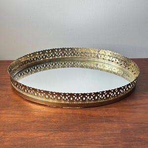 Puede incluir: Una bandeja ovalada con una base de espejo reflectante y un borde decorativo de metal en tono dorado. El borde elevado presenta un patrón calado repetitivo, proporcionando un marco estructurado alrededor de la superficie de vidrio.