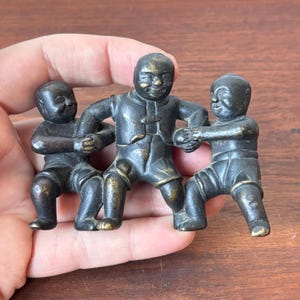 May include: A bronze figurine of three figures in a playful pose. The figures are dressed in traditional clothing and appear to be engaged in a game or activity. The figurine has a weathered appearance.