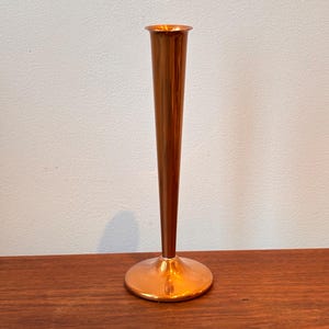 May include: A copper-colored vase with a flared top and a slender, conical body. The vase has a circular base and stands on a wooden surface. The vase is likely used for displaying flowers or as a decorative item.