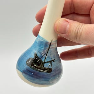 May include: A ceramic spoon with a white handle and a blue painted bowl. The bowl features a black sailboat design against a blue and white background, evoking a nautical theme. The spoon is held against a white background.
