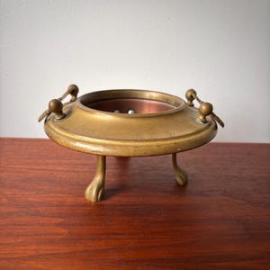 May include: An antique brass incense burner. It features a circular design with three claw feet and a copper interior. The burner has two decorative handles. The brass has a warm, aged patina, indicating its vintage origin.
