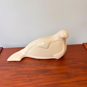 May include: A cream-colored ceramic walrus figurine with two small, copper-colored tusks. The walrus is in a resting position, with its flippers tucked in. The figurine is on a dark wood surface.