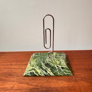 May include: A green marble paperclip holder with a silver-coloured paperclip. The marble base is rectangular with a polished finish, displaying a mix of green and white veining. The paperclip is upright, creating a functional and decorative desk accessory.