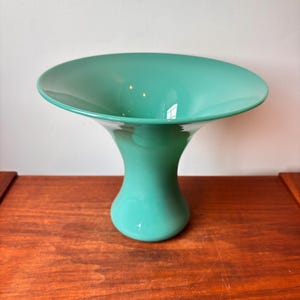 Può includere: Un vaso di vetro turchese con una coppa ampia e poco profonda e una base svasata. Il vaso ha una finitura liscia e lucida e poggia su una superficie di legno. Il design è moderno ed elegante, adatto per esporre fiori o come elemento decorativo.