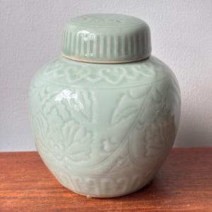 May include: A pale green ceramic ginger jar with a matching lid. The jar features a glossy finish and embossed floral patterns around the body, with a decorative border near the top and bottom. The lid has a ribbed edge and a smooth, rounded top.