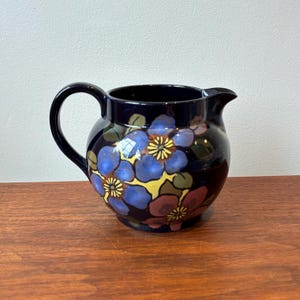 Op de afbeelding: Een donkerblauwe keramische kan met een afgeronde body en een handvat. De kan is versierd met blauwe, gele en kastanjebruine bloemmotieven. De kan heeft een tuit om uit te schenken.