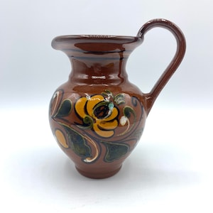 May include: A brown ceramic jug with a floral design in yellow, green, and black. The jug has a wide mouth and a handle.