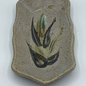 May include: A small, rectangular ceramic dish with a speckled glaze. The dish has a wavy edge and features a hand-painted design of a stylised flower in shades of brown, green, and blue.