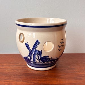 Peut inclure: Pot en céramique crème avec un motif de moulin à vent bleu et des découpes circulaires. Le récipient a un bord bleu et un motif bleu de moulin à vent, d'arbres et d'oiseaux en vol.