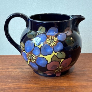 Op de afbeelding: Een donkerblauwe keramische kan met een afgeronde body en een gebogen handvat. De kan is versierd met blauwe, gele en roze bloemmotieven en groene bladeren. De kan heeft een tuit om uit te schenken.