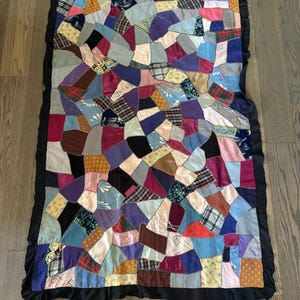 May include: A rectangular crazy quilt featuring an irregular patchwork of multicoloured fabric scraps in various textures and patterns, including plaid, floral, and solid colours. The quilt is finished with a solid black ruffled fabric border.