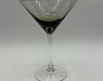 May include: A clear martini glass with a gray-tinted bowl and a long, slender stem. The bowl has a decorative pattern of small, etched circles. The glass sits on a round base, and is set against a white background.