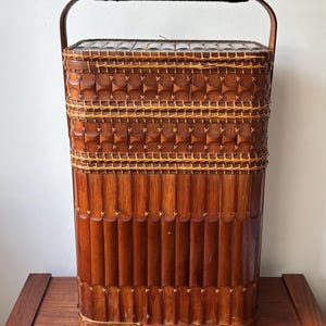May include: A tall, rectangular brown bamboo basket with a lid and a high arched handle. The exterior features vertical bamboo slats, horizontal woven bands, and decorative cross-stitching in a lighter tan color throughout the structure.