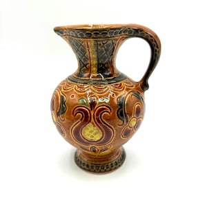 May include: A small, decorative ceramic pitcher with a handle and spout. The pitcher is primarily brown with intricate floral and geometric patterns in shades of green, yellow, and red. The base is a small, rounded foot.