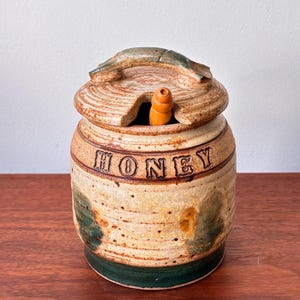 Peut inclure: Un pot à miel en céramique fait à la main avec un couvercle et un bâtonnet à miel en bois. Le pot est de couleur beige avec des accents marron et vert, et le mot "HONEY" est inscrit au milieu. Le couvercle a une poignée.
