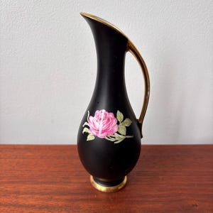 Könnte beinhalten: Schwarze Keramikvase mit einem rosa Rosenmuster. Die Vase hat einen goldenen Griff und Zierleisten. Die Rose ist mit rosa Blütenblättern und grünen Blättern bemalt. Die Vase ist etwa 15 cm hoch.
