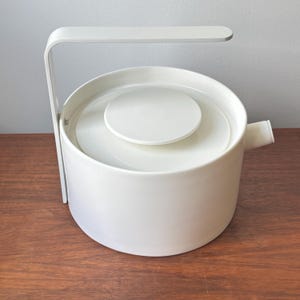 May include: A white ceramic teapot with a cylindrical body, a flat circular lid, and a single angular handle that extends vertically from the side and arches over the top. A small, short spout is positioned on the opposite side of the handle.