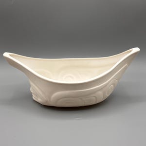 Peut inclure: Un bol en céramique blanche avec un motif sculpté. Le bol est en forme de canoë et a une surface lisse et arrondie.