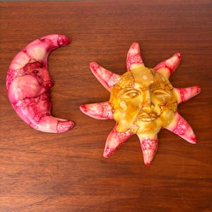 May include: Decorative sun and moon wall art. The sun has a golden face with red and white accents. The crescent moon is pink with white accents. Both are placed on a dark brown wooden surface.