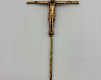 May include: A bronze-colored crucifix with a detailed figure of Jesus Christ. The cross has horizontal arms and a vertical base with a decorative pattern. The figure is in a traditional pose, arms outstretched.