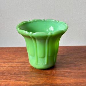 Op de afbeelding: Een lichtgroene glazen vaas met een brede, uitlopende rand met een geschulpte afwerking en verticale ribbeldetails langs het lichaam. De vaas heeft een gladde, glanzende afwerking en een afgeronde basis in een consistente jadegroene kleur.