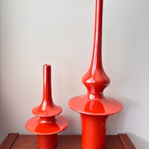 May include: Two vibrant red glass vases. The taller vase has a flared base, a bulbous midsection, and a long, slender neck. The shorter vase has a tiered design with a cylindrical base and a short, straight neck. Both vases are glossy.