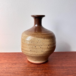 May include: A small, handcrafted ceramic vase with a rounded body and a narrow neck. The vase features a speckled, tan-colored exterior with a darker brown rim and neck. The vase is a decorative item.