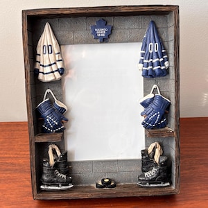 May include: A decorative photo frame with a hockey theme. The frame features miniature hockey jerseys, gloves, and skates. The frame is brown with a gray brick-like background and the text "TORONTO MAPLE LEAFS".