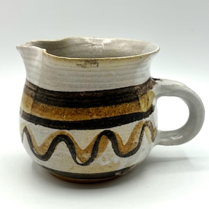 May include: A white ceramic jug with a brown and yellow striped design and wavy lines. The jug has a handle and a rounded body.