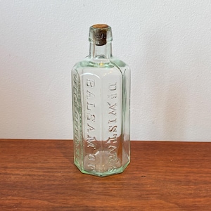 May include: A clear, rectangular glass bottle with beveled edges and a cork stopper. The glass is embossed with the text DR. WISTAR'S BALSAM OF WILD CHERRY. The bottle features a narrow neck and a flat base, showcasing a vintage medicinal design.