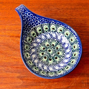 May include: A blue and white ceramic spoon rest with a peacock feather design. The spoon rest has a teardrop shape with a handle on one end. The interior features a pattern of green and white circles and black dots.