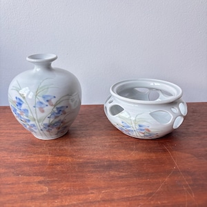 May include: Two ceramic vases with floral designs. One is a small, round vase with a narrow neck. The other is a small, round bowl with cut-out designs. Both are painted with blue and pink flowers and green stems.