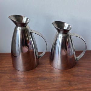 May include: Two stainless steel pitchers with a polished, reflective finish. Each pitcher features a tapered, conical body, a wide pouring spout, and a curved handle attached to the side. The pitchers are of different heights and widths.