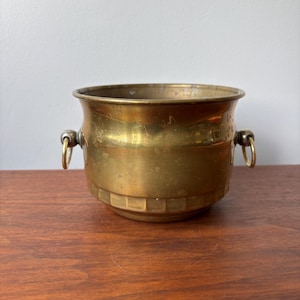 May include: A vintage brass planter with a rounded body and two ring handles. The planter has a decorative band near the base and a slightly flared rim. The brass has a warm, aged patina, suggesting its antique nature. The planter is suitable for indoor or outdoor use.
