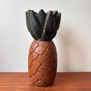 May include: A carved wooden pineapple sculpture. The pineapple has a brown body with a textured, scale-like pattern and a dark brown, spiky top. The sculpture is displayed on a wooden surface.