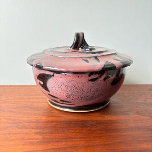 Könnte beinhalten: Eine Keramikschale mit Deckel und einem rosa-schwarzen, gesprenkelten Design. Die Schale hat eine runde Form mit Deckel und Griff. Deckel und Schale sind mit schwarzen, abstrakten Formen verziert. Die Schale steht auf einer Holzoberfläche.