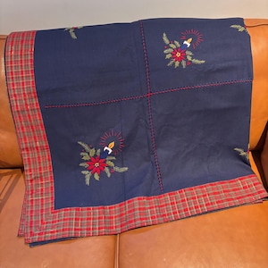 Puede incluir: Un mantel navideño azul marino con ribete a cuadros rojos. El mantel presenta bordados de flores de pascua, velas y vegetación. Las costuras rojas acentúan las costuras y los detalles bordados. El mantel está doblado sobre una superficie de cuero marrón.