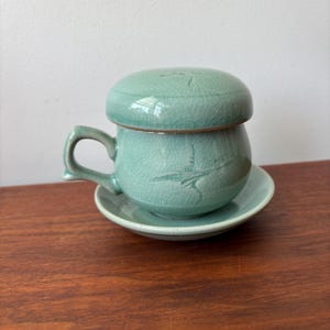 Könnte beinhalten: Ein Tee-Service aus Keramik in Celadon-Farbe, bestehend aus einer Tasse mit Henkel, Deckel und Untertasse. Das hellgrüne Set weist geätzte Designs auf. Auf einer Holzoberfläche platziert.